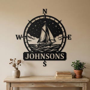 Personalized nautical compass metal wall decor featuring a sailboat at sea with custom family name, designed in a round coastal style. Available in multiple sizes and finishes including black, gold, silver, bronze, and white. Ideal for living rooms, entryways, beach houses, lake houses, or outdoor walls. Easy to hang and weather resistant. A unique gift for sailors, ocean lovers, housewarming, weddings, or modern coastal home decor.