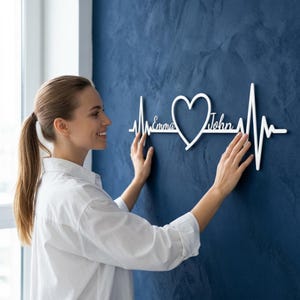 May include: A white metal wall decoration in the form of a heartbeat, featuring a heart shape. The names "Emma" and "John" are inscribed. The artwork is displayed against a dark blue wall.