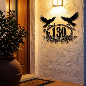 Puede incluir: Letrero de número de casa de metal negro con el número 130 en un marco ovalado decorativo, adornado con hojas y dos siluetas de pájaros. El letrero está montado en una pared blanca texturizada, iluminada por una luz cálida.
