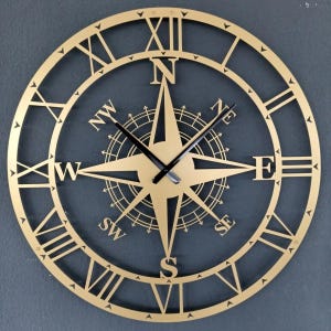 Könnte beinhalten: Eine kreisförmige, goldfarbene Wanduhr mit einem Kompassrosen-Design in der Mitte, das die Himmelsrichtungen N, S, O, W, NW, NO, SW und SO zeigt. Der äußere Ring zeigt römische Ziffern von I bis XII und zwei schwarze Uhrzeiger in der Mitte.