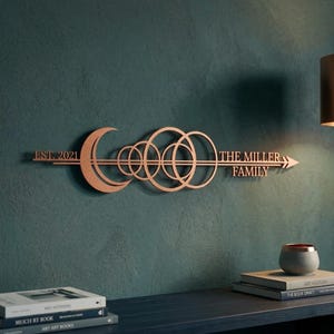 Puede incluir: Una pieza de arte mural de metal color cobre con el texto "EST. 2021" y "THE MILLER FAMILY". El diseño incluye una luna creciente, círculos superpuestos y una flecha. La obra de arte está montada en una pared verde azulado.