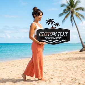 May include: A black metal sign with the words "CUSTOM TEXT - BEACH HOUSE" in white and copper lettering, featuring palm tree silhouettes. The sign is held on a beach with a blue ocean and palm tree in the background.