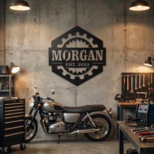 Puede incluir: Un letrero de metal negro con el nombre "MORGAN" y "EST. 2025" está montado en una pared de hormigón. El letrero tiene un diseño en forma de engranaje. Una motocicleta plateada está estacionada debajo del letrero. La escena está ambientada en un garaje o taller.