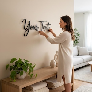 Puede incluir: Una persona está montando un letrero de pared negro con la frase "Your Text" en una pared beige. El letrero está hecho de un material oscuro. La habitación tiene una estética moderna con una consola de madera y un sofá.