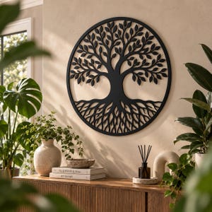 May include: A circular black metal wall art piece featuring a detailed tree of life design with intricate branches, leaves, and roots enclosed within a solid ring frame, mounted on a plain wall above a wooden cabinet with various decorative items.