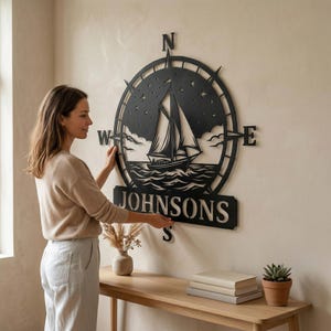 Personalized nautical compass metal wall decor featuring a sailboat at sea with custom family name, designed in a round coastal style. Available in multiple sizes and finishes including black, gold, silver, bronze, and white. Ideal for living rooms, entryways, beach houses, lake houses, or outdoor walls. Easy to hang and weather resistant. A unique gift for sailors, ocean lovers, housewarming, weddings, or modern coastal home decor.