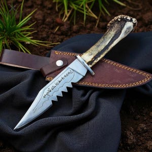 May include: A hunting knife with a serrated blade and a bone handle rests in a brown leather sheath. The blade has etched text. The sheath has a leather belt loop and a snap closure. The knife is on a black cloth.