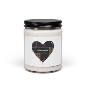 May include: A white candle jar with a black lid. The label features a heart-shaped patchwork design in red, green, and yellow plaid, secured with safety pins. The text "GRUNGE REVIVAL" is printed on the label.