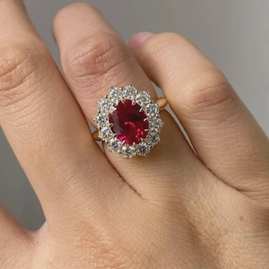 Puede incluir: Un anillo de oro con una gran gema roja oscura de corte ovalado, rodeada por un halo de gemas claras más pequeñas. El anillo está engastado en una banda de oro, creando un diseño clásico y elegante.