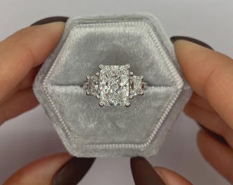 May include: A radiant-cut diamond ring with side stones, set in a silver band, rests in a gray velvet ring box. The ring features a large, rectangular center stone flanked by two smaller stones, creating a classic and elegant design.
