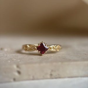 May include: A gold ring with a square, deep red gemstone at the center. The band features intricate floral detailing. The ring is set against a neutral background, highlighting its elegant design.