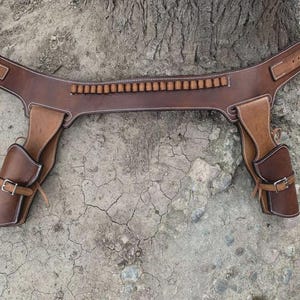 Peut inclure: Une ceinture de pistolet en cuir marron avec deux étuis et une rangée de boucles de balles. La ceinture a une fermeture à boucle et est cousue avec du fil blanc. Les étuis ont des fermetures à boucle.