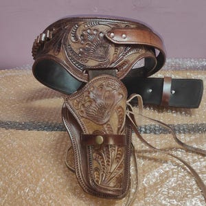 Op de afbeelding: Een bruin leren pistoolholster en riemset met ingewikkelde bloemmotieven. De riem heeft patronenlussen en een gesp. Het holster is ontworpen voor een revolver. Afmetingen in cm.