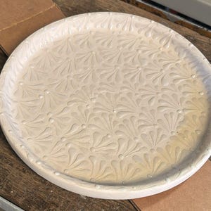 Puede incluir: Una bandeja redonda de cerámica blanquecina con un patrón floral en relieve repetido. La bandeja tiene un borde ligeramente elevado y se encuentra sobre una superficie de madera. Esta bandeja decorativa es adecuada para servir o exhibir.