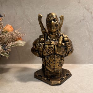 Puede incluir: Busto dorado de un personaje de superhéroe, con una cara enmascarada y dos espadas en la espalda. La figura lleva un traje blindado detallado. El busto está montado sobre una base cuadrada, mostrando un diseño detallado.