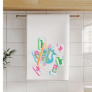 May include: White hand towel with a colorful peacock and Mahjong tile design. The peacock is blue with a floral design. The Mahjong tiles are pink, green, and orange. The towel is hanging on a silver bar.