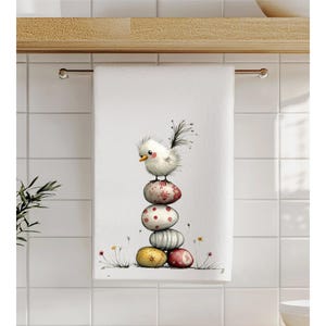 May include: White hand towel featuring a whimsical illustration of a fluffy white bird perched atop a stack of colorful Easter eggs. The eggs are decorated with floral and polka dot patterns. The towel is hanging on a metal rod.