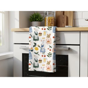 May include: A white kitchen towel featuring a repeating pattern of illustrated mice wearing aprons, cupcakes, cherries, lemons, and pitchers. The mice are depicted in various colors including white, tan, and light gray, with blue and green accents.