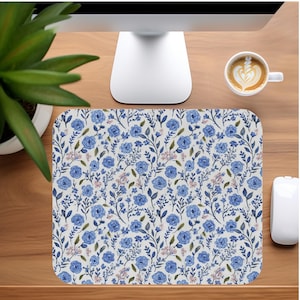 Puede incluir: Una alfombrilla de ratón floral con un patrón de rosas azules y hojas verdes sobre un fondo blanco. La alfombrilla es rectangular y está sobre un escritorio de madera, junto a un ratón de ordenador blanco y una taza de café.