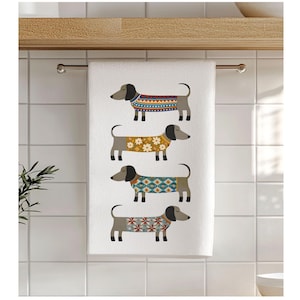 Peut inclure: Une serviette à main rectangulaire blanche ornée d'un motif vertical répétitif représentant quatre chiens teckels. Chaque chien est illustré de profil, portant un pull coloré différent avec des motifs géométriques, floraux et en losange.