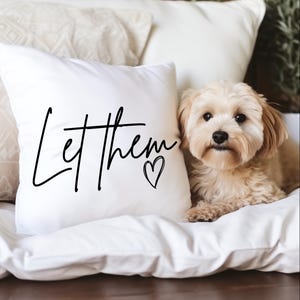 Puede incluir: Un cojín cuadrado blanco con las palabras "Let them" en escritura negra y un símbolo de corazón. Un perro pequeño, de color marrón claro, está acostado junto al cojín sobre una superficie blanca.