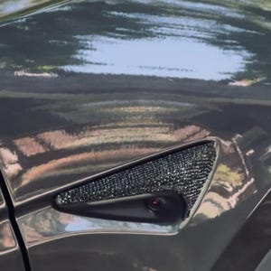 May include: Close-up of a dark gray car panel with a triangular vent covered in small, sparkling black embellishments. The panel reflects the sky and surrounding environment, creating a glossy appearance. A small red light is visible.