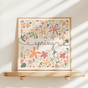 May include: A framed floral art print with the word "spring" in cursive. The artwork features a variety of colorful flowers in shades of pink, orange, yellow, and green. The frame is a light wood color, and the print is displayed on a wooden shelf.