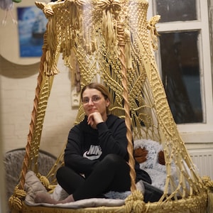 Könnte beinhalten: Ein beigefarbener Makramee-Hängesessel mit dekorativem Baldachin. Der Stuhl ist an der Decke aufgehängt und hat ein gewebtes Design. Die Person trägt einen schwarzen Kapuzenpullover und eine schwarze Hose.