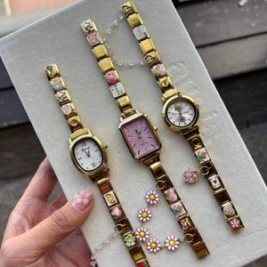 Puede incluir: Tres relojes dorados con pulseras de abalorios decorativas. Uno tiene una esfera ovalada blanca, otro una esfera rectangular rosa y el tercero una esfera redonda blanca. Las pulseras presentan abalorios coloridos y detalles florales.