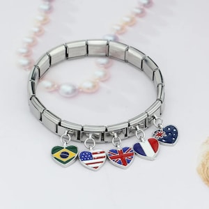 Könnte beinhalten: Ein silberfarbenes Charm-Armband mit herzförmigen Anhängern. Jeder Anhänger zeigt die Flagge eines Landes, darunter Brasilien, die USA, Großbritannien, Frankreich und Australien. Das Armband wird vor einem hellen Hintergrund gezeigt.