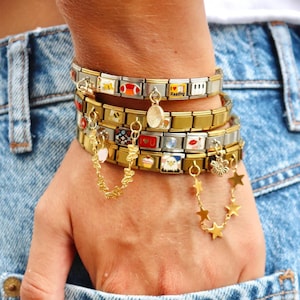 May include: Several gold and silver charm bracelets are stacked on a wrist. The bracelets feature various charms, including a heart, a star, and the word "Reading". The bracelets are accessorized with dangling charms and are set against a denim background.