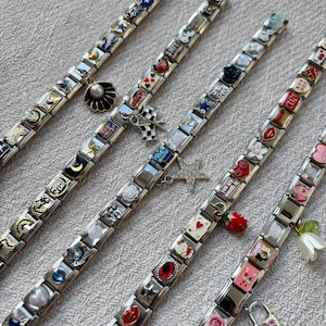 May include: Several silver charm bracelets are shown, each with a variety of decorative charms. The charms feature hearts, stars, flowers, and other designs, some with colourful enamel. The bracelets are displayed on a light-coloured surface.
