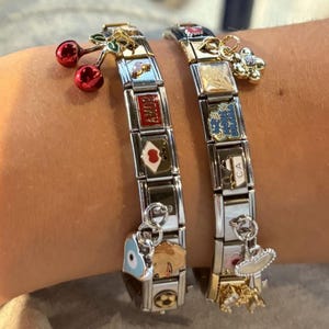 Op de afbeelding: Twee bedelarmbanden met zilveren schakels en gouden accenten. Eén armband heeft rode kersenbedels, terwijl de andere een bloembedel heeft. Beide armbanden hebben diverse decoratieve bedels met kleurrijke emaille ontwerpen.