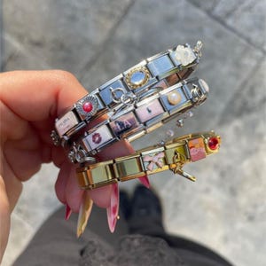 Könnte beinhalten: Drei Charm-Armbänder mit verschiedenen dekorativen Charms. Die Armbänder sind silber- und goldfarben. Zu den Charms gehören ein roter Edelstein, eine Perle und Charms mit den Prada- und LA-Logos. Die Armbänder werden in einer Hand gehalten.