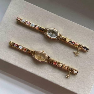 Puede incluir: Dos relojes de pulsera dorados con esferas ovaladas y correas adornadas con dijes. Las correas presentan dise&ntilde;os en miniatura coloridos, incluyendo flores, corazones y un letrero de la Ruta 66. Cada reloj tiene un peque&ntilde;o dije colgante.