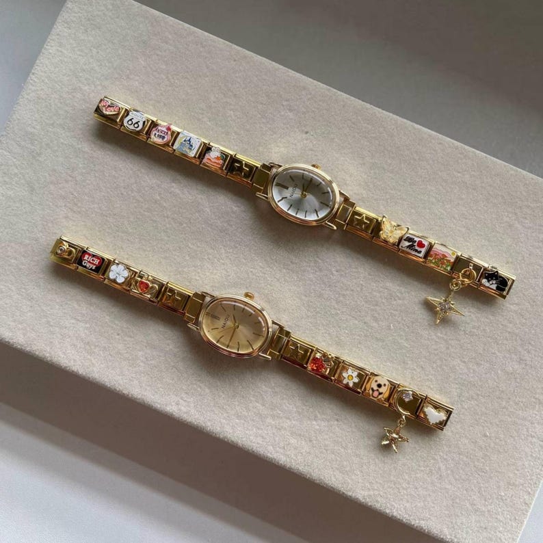 Puede incluir: Dos relojes de pulsera dorados con esferas ovaladas y correas adornadas con dijes. Las correas presentan dise&ntilde;os en miniatura coloridos, incluyendo flores, corazones y un letrero de la Ruta 66. Cada reloj tiene un peque&ntilde;o dije colgante.