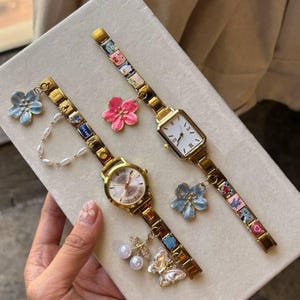 Puede incluir: Dos relojes de pulsera dorados con dijes decorativos. Uno tiene una esfera redonda, el otro rectangular. Ambos tienen correas de eslabones dorados con detalles coloridos y dijes de flores. Uno tiene una cadena de perlas y un dije de mariposa.