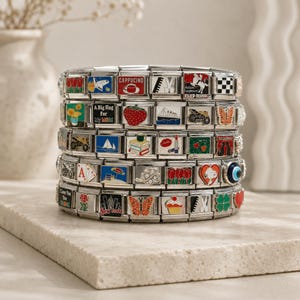 Puede incluir: Una pulsera de eslabones modulares en tono plateado con una variedad de dijes de esmalte coloridos, que incluyen imágenes de flores, una fresa, un cupcake, una mariposa, un velero, una taza de café y textos como New York y A Big Hug for My MOM.