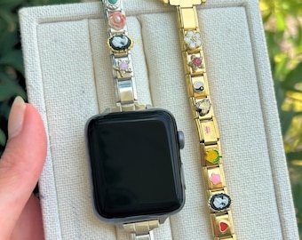 Op de afbeelding: Een Apple Watch met twee decoratieve bandjes. Eén bandje is zilverkleurig met kleurrijke bedels, de andere goudkleurig met diverse bedels. De wijzerplaat van het horloge is zwart.