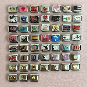 Puede incluir: Colección de abalorios plateados con varios diseños, incluyendo corazones, banderas y textos como "CRAZY" y "USA". Algunos abalorios presentan imágenes de un perro, un Mickey Mouse y un mando de videojuegos. Los abalorios están dispuestos sobre una superficie rosa.