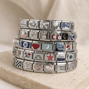 Puede incluir: Una pila de cinco pulseras modulares de tono plateado con varios eslabones de esmalte y metal. Los diseños incluyen una corona, un lazo, cerezas, un ojo turco, una fresa, una estrella, un gato y textos como Princess, Be Mine, Make A Wish y Bling Bling.