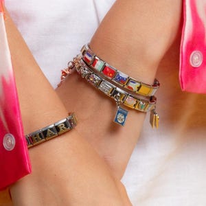 Puede incluir: Se muestran varias pulseras de abalorios, con varios abalorios. Una pulsera tiene abalorios con temas de banderas, mientras que otra tiene abalorios con las letras "HARI". Las pulseras son plateadas y doradas, con acentos coloridos.