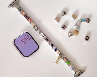 Op de afbeelding: Zilveren horlogeband met meerdere vierkante bedels met verschillende ontwerpen en kleuren. Een goudkleurige bedel hangt aan de band. Een paarse wijzerplaat toont de tijd. Extra bedels liggen in de buurt verspreid.