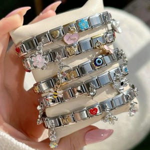 May include: Several stainless steel charm bracelets with various decorative charms. Charms include a pink heart with "Love" text, a blue flower, a bee, and a red heart. The bracelets are stacked on a cream-colored surface.