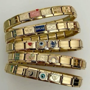 May include: Gold-coloured charm bracelets with various decorative charms. Charms include a butterfly, heart, eye, and the word "DREAM". The bracelets are stacked and displayed on a white surface.