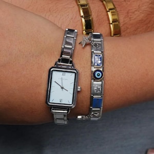 Puede incluir: Reloj rectangular plateado con esfera blanca y números romanos. Una pulsera de abalorios con dijes de estrella, azules y de ojo turco está al lado del reloj. Una pulsera dorada está en el fondo.