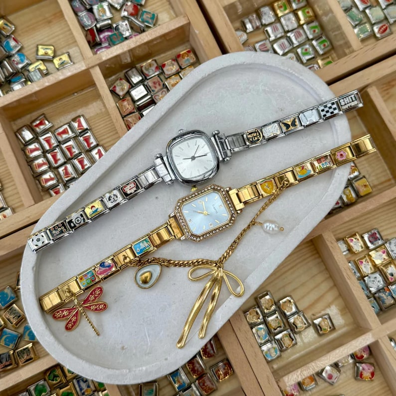 May include: Two square-faced watches with decorative charm bracelets. One watch is silver-toned with a white face, the other is gold-toned with a light blue face and a diamond-like border. A gold-toned charm and bow pendant are attached.