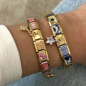 Könnte beinhalten: Zwei goldfarbene Charm-Armbänder mit bunten Emaille-Kacheln und Stern-Anhängern. Ein Armband zeigt das Wort "IBIZA" und rosa Kacheln. Das andere hat blaue und weiße Kacheln. Beide werden am Handgelenk getragen.