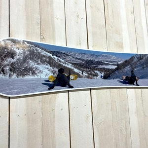 Puede incluir: Una tabla de snowboard blanca con una imagen colorida de una escena de monta&ntilde;a nevada con dos personas haciendo snowboard cuesta abajo. La imagen est&aacute; impresa en la parte superior de la tabla de snowboard.