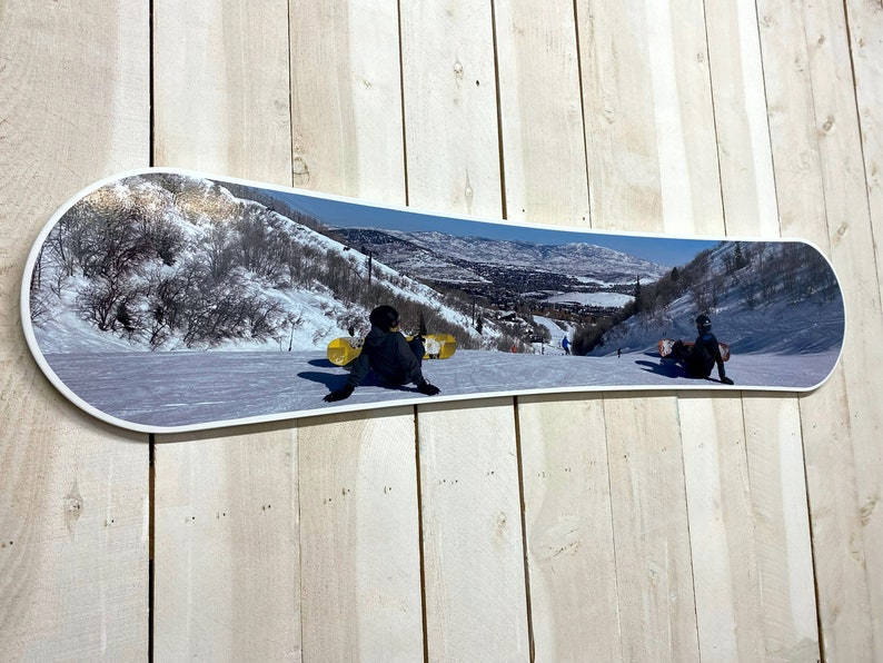Puede incluir: Una tabla de snowboard blanca con una imagen colorida de una escena de monta&ntilde;a nevada con dos personas haciendo snowboard cuesta abajo. La imagen est&aacute; impresa en la parte superior de la tabla de snowboard.